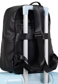 Sac à dos noir texturé fixé à la poignée d'une valise bleue claire avec des sangles noires ajustables. Il possède une poche zippée à l'arrière.