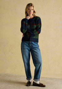 Pletený tmavomodrý cardigan so zelenými a červenými geometrickými vzormi, s okrúhlym výstrihom a zapínaním na gombíky. Spárovaný so svetlomodrými džínsami a hnedými mokasínami.