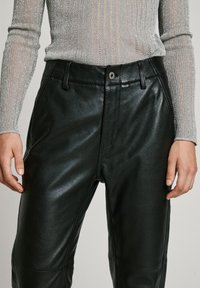 Black leather pants with a smooth texture, front zipper, and button closure. Worn with a fitted, lightly ribbed gray top.