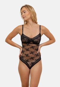 Bodysuit en dentelle noire avec un tissu transparent, des motifs floraux, des bretelles réglables, et un design à bord ondulé au niveau du décolleté et des ouvertures des jambes.