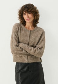 Bruine, gestructureerde cardigan met lange mouwen en een ronde hals. Voorzien van een knoopsluiting en een licht cropped ontwerp. Gecombineerd met een zwarte rok.