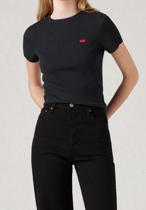 Kobieta w dopasowanej czarnej koszulce z krótkim rękawem z małym czerwonym logo Levi's i czarnych spodniach z wysokim stanem na jednolitym tle.