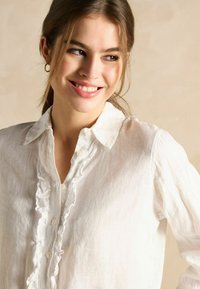 Biely ľanový košeľový top so zúbkovaným zapínaním na gombíky a golierom, s jemnou, mierne priesvitnou textúrou.