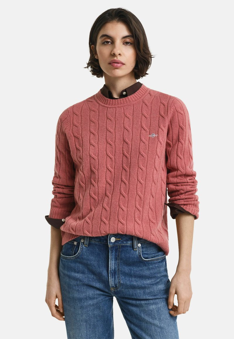 Kabelstrickpullover in staubigem Rosa, runder Ausschnitt, aufgeschlagene Ärmel, über einem dunklen Kragenhemd getragen. Kombiniert mit blauen Denim-Jeans.