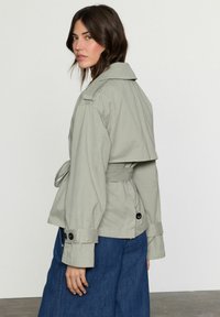 Giacca grigia chiara cropped con vita cintata, maniche larghe e grandi bottoni neri, indossata su una gonna di jeans.