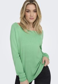 Groene trui met lange mouwen van zacht gebreid materiaal, met een relaxte pasvorm en een bateauhals, gecombineerd met donkere jeans. Accent details omvatten geribbelde manchetten.