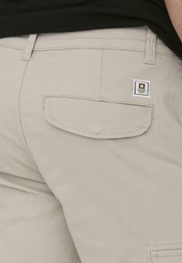 Pantalones cargo beige con un bolsillo trasero que presenta un pequeño parche cuadrado de la marca. El material parece duradero con una textura suave.