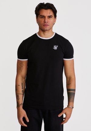 Mann mit dunklem Haar, trägt ein schwarzes T-Shirt mit weißem Rand und Logo auf der Brust, zeigt verschiedene Arm-Tattoos, steht vor einfachem Hintergrund.