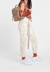 Sac à dos brun tacheté, haut à manches longues orange à motifs, pantalon beige clair tie-dye, et baskets blanches avec des chaussettes orange à la cheville.