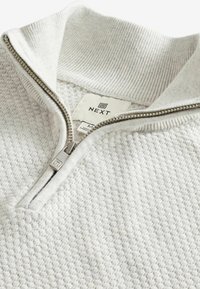 Pull en maille texturée gris clair avec fermeture éclair, col côtelé et finition lisse. L'étiquette indique la marque "NEXT" et la taille "M".