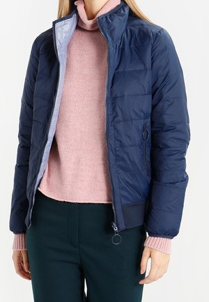 Mujer con una chaqueta acolchada azul marino sobre un suéter de cuello alto rosa y pantalones oscuros, de pie frente a un fondo liso.