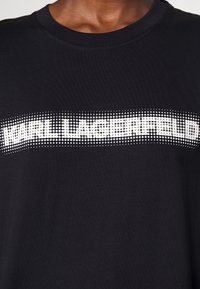 Camiseta de algodón negra con cuello redondo, que presenta un gráfico de texto en blanco con puntos que dice "KARL LAGERFELD" en el pecho.