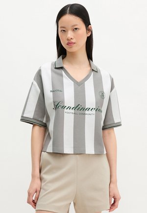 Νεαρή γυναίκα με μακριά μαύρα μαλλιά που φορά γκρι και λευκή ριγέ πόλο μπλούζα με το κείμενο «Scandinavian Football Community» και μπεζ σορτς.