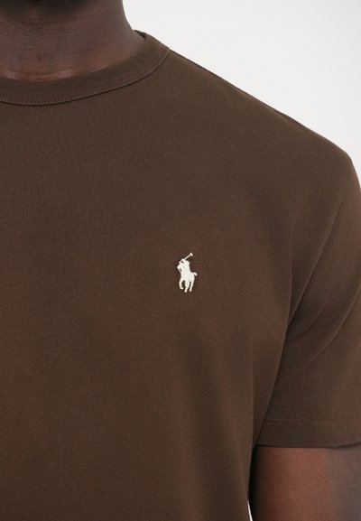 Barna pamut póló kerek nyakkivágással. A bal mellrészen kis fehér pólójátékos logó hímzéssel. Sima textúrájú.
