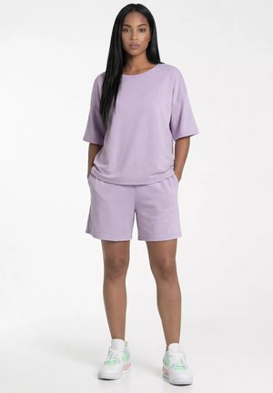 Kobieta stoi z rękami w kieszeniach, ubrana w luźny liliowy top z krótkim rękawem, pasujące szorty oraz białe trampki z pastelowymi akcentami.