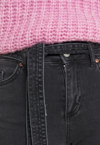 Los jeans de mezclilla negros tienen un cinturón y un bolsillo delantero con remaches de cobre. Un suéter de punto grueso rosa asoma por encima de la cintura.