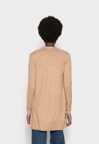 Cardigan beige à manches longues en tissu doux, avec un col rond et des poignets côtelés, offrant une coupe décontractée et légèrement allongée.