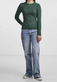 Femme portant un pull côtelé vert foncé ajusté, un jean large bleu clair et des baskets basses noires, debout devant un fond uni.