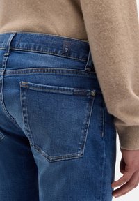 Niebieskie dżinsy z denimu mają średni stan, pięciokieszeniowy fason i delikatne przetarcia, zestawione z beżowym swetrem dzianym.