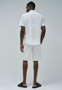 Camisa blanca de lino de manga corta con cuello, combinada con pantalones cortos blancos a juego. Lleva sandalias negras sin hebilla, de pie frente a un fondo sencillo.