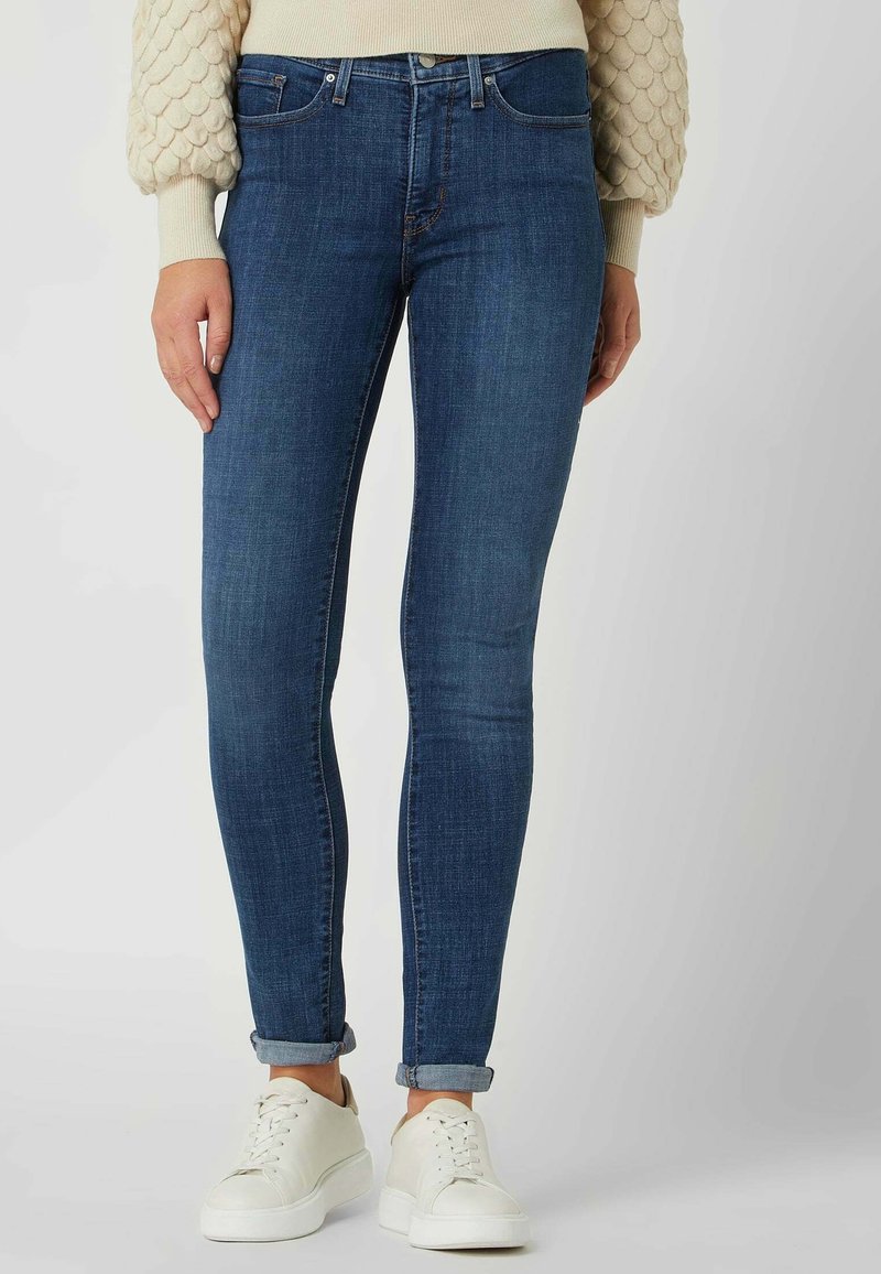 Personne portant un jean skinny bleu foncé retroussé, un pull texturé crème et des baskets blanches, debout sur un fond gris clair uni.