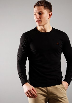 Maglia nera a maniche lunghe con collo rotondo e design aderente. Presenta un piccolo logo arancione sul lato sinistro del petto. Texture liscia, dettagli minimi.