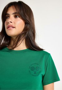 Grøn bomulds-t-shirt med rund halsudskæring og korte ærmer, med et tennislogo trykt i sort på venstre bryst.