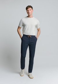 Lichtgrijze crewneck t-shirt, marineblauwe broek met elastische taille en bruine trekkoord, en beige sneakers; casual silhouet met gladde texturen.