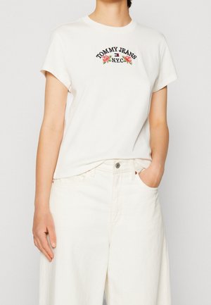 Nainen pukeutunut valkoiseen Tommy Jeans -t-paitaan, jossa on kukkakirjailu, ja valkoisiin korkeavyötäröisiin housuihin, toinen käsi taskussa.