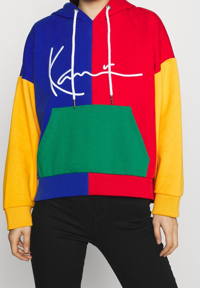 Personne portant un sweat à capuche multicolore avec des sections bleues, rouges, jaunes et vertes et des cordons blancs, associé à un pantalon noir.