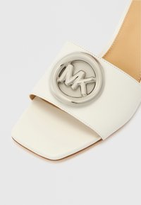 Bijela kožna natikača s kvadratnim vrhom, s velikim okruglim srebrnim logotipom MK na remenu. Mekani bež uložak.