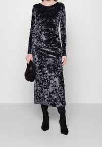 Robe longue en velours noir avec manches longues, col rond et silhouette ajustée. Portée avec des bottes noires montantes jusqu'aux genoux et une pochette texturée.