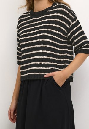 Žena nosící černo-bílý pruhovaný pletený top s krátkým rukávem, má ruku v kapse černé sukně nebo kalhot.