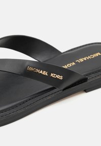 Sandal in pelle nera con cinturini larghi. Presenta il logo dorato "MICHAEL KORS" su un cinturino. Suola piatta, design minimalista, texture liscia.