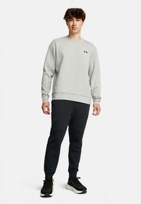 Sweatshirt de tecido cinzento com punhos canelados, combinado com calças de treino pretas afuniladas com detalhes em bolsos laterais. Ténis desportivos pretos com solas brancas.