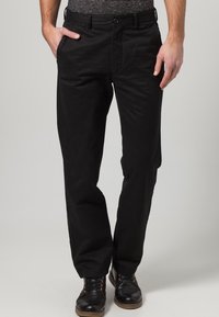 Pantaloni di cotone neri con design a gamba dritta, dotati di tasche laterali, chiusura a bottone e una texture sottile. Abbinati a scarpe scure allacciate.