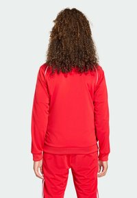 Κόκκινη αθλητική φόρμα με ομαλή υφή, με λευκές ρίγες στα μανίκια και εφαρμοστό σχεδιασμό. Εμφανίζεται η πίσω όψη.