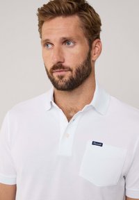Biała koszulka polo z teksturowanej tkaniny, z kołnierzykiem, trzema guzikami i kieszonką na piersi z granatową metką.