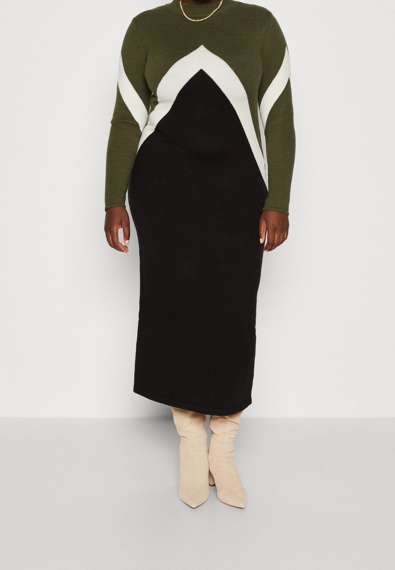 Personne portant une robe à manches longues verte, noire et blanche avec un motif géométrique et des bottes beige mi-mollet sur un fond uni.