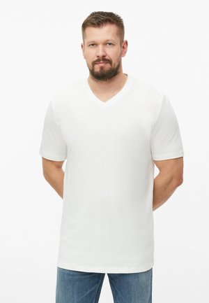 Man met kort bruin haar en baard staat naar voren gericht, draagt een eenvoudig wit V-hals T-shirt en blauwe jeans tegen een witte achtergrond.