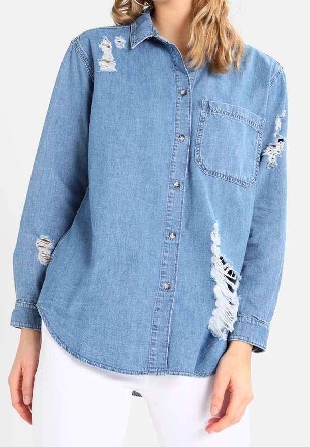 Chemise en jean bleu clair avec des déchirures usées au niveau du col, de la manche et de la poche avant, portée avec un pantalon blanc.