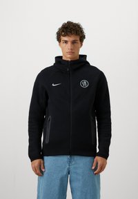 Sort hættejakke med lynlås lavet af tekstureret stof, med et Nike-logo og et rundt emblem på brystet. To sidelommer med lynlås.