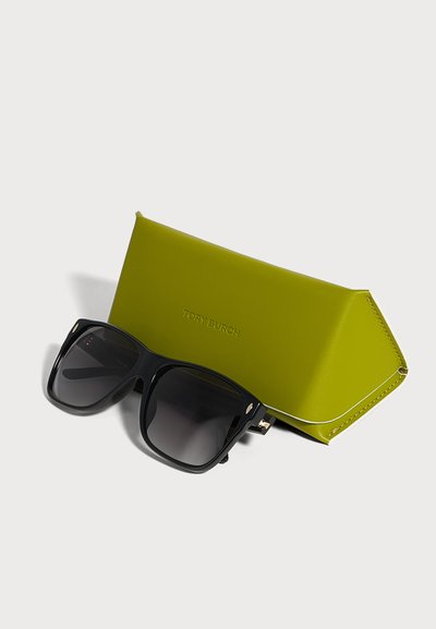 Des lunettes de soleil noires oversize aux verres foncés sont posées contre un étui Tory Burch vert citron, sur un fond clair.