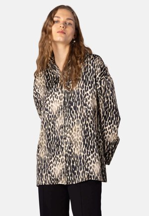 Ung kvinde med langt bølget hår iført en løs skjorte med leopardprint og sorte bukser, står foran en simpel lys baggrund.