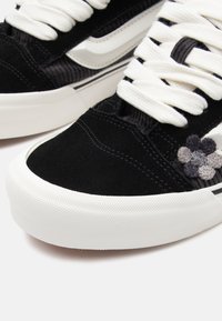 Black suede sneaker with white rubber sole, features textured accents, white laces, and embroidered floral detail in shades of gray.