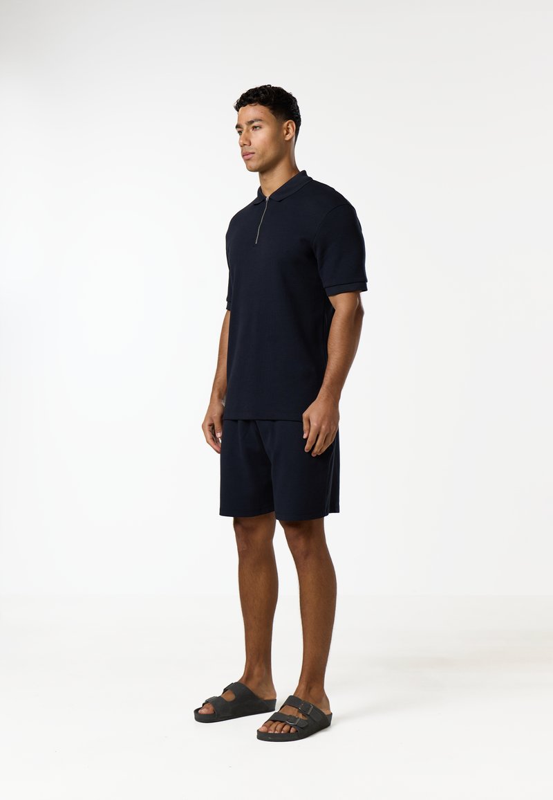 Homme debout portant un polo noir à manches courtes avec fermeture éclair, un short noir et des sandales à brides noires sur un fond blanc uni.