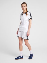 Hvid atletisk polo shirt med sort chevron-detaljering på ærmerne, parret med hvide shorts, blå sko og hvide ankelstrømper.