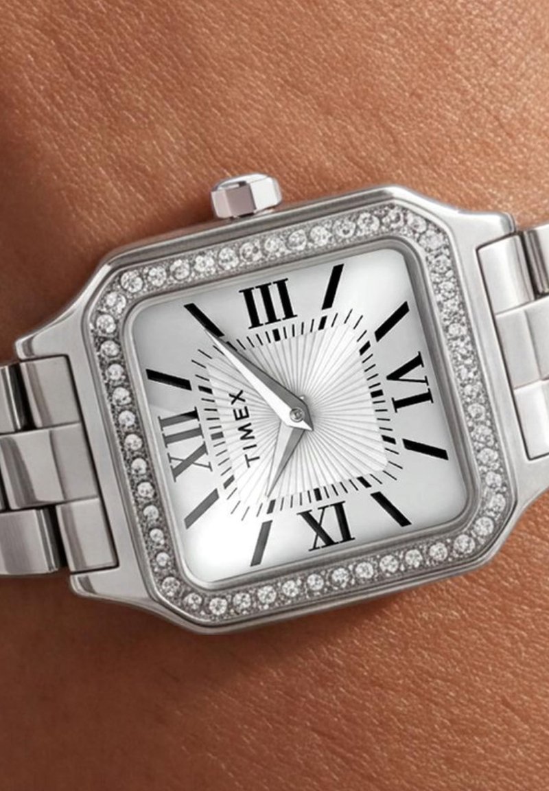 Ασημένιο ρολόι χειρός Timex με τετράγωνο καντράν, ρωμαϊκούς αριθμούς, στεφάνη με κρύσταλλα και μεταλλικό μπρασελέ σε καφέ επιδερμίδα.