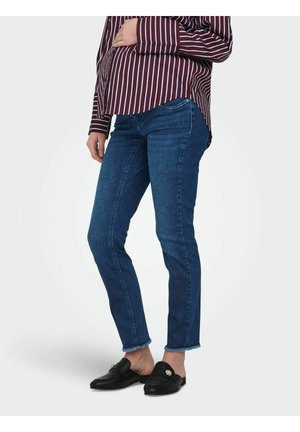 Person som har på seg mørkeblå, smale jeans med frynsete kanter, svarte slip-on loafers og en burgunderrød langermet skjorte med striper, stående med den ene hånden på hoften.