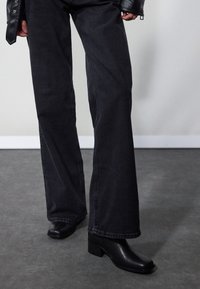 Personne portant un jean noir à jambes larges et des bottes noires à talons carrés, debout sur un sol gris contre un mur clair.